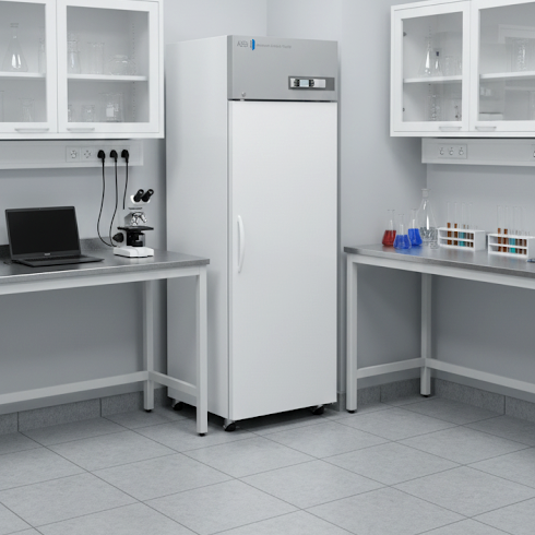 A laboratory with gray tile flooring, white walls, and white cabinets. In the center is a tall, white laboratory refrigerator. Counters on either side hold a laptop, a microscope, and several racks of glass test tubes containing colored liquids.
