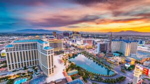 Aerial view of the Las Vegas Strip at sunset, showing bright hotel lights, casinos, the Bellagio fountains in the foreground, and the replica Eiffel Tower. Colorful sky with clouds and distant mountains frame the bustling cityscape.