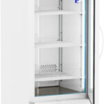 An upright, white laboratory refrigerator with a glass door is open, revealing three empty shelves. The digital display on top shows the internal temperature. The refrigerator is on wheels and has a vented panel at the bottom.