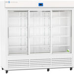 A large, commercial white refrigerator with three sliding glass doors and multiple empty wire shelves. A digital temperature display is above the center door. Only one green tennis ball sits on the bottom right shelf; the rest of the shelves are empty.
