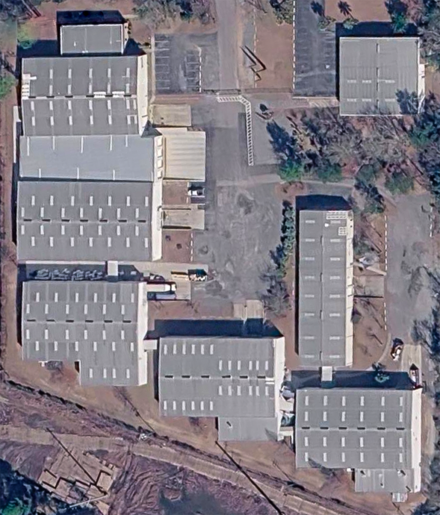 Aerial view of a large industrial facility with multiple rectangular buildings, gray roofs, parking lots, scattered cars, and surrounding trees. The area features paved roads and unpaved ground nearby. The overall scene is orderly and functional.