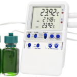 A digital thermometer displays temperatures on a large screen, showing readings of 23.92°C and 21.48°C. Two green liquid-filled vials with teal caps are in front, and two white sensor cables are plugged into the device’s right side.
