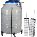 A large silver cryogenic storage tank labeled American BioTech Supply with a blue lid and attached gauges stands on wheels. Next to it are two vertical racks with multiple drawers and a black measuring stick, used for storing biological samples.