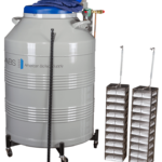 A large, gray cryogenic storage tank labeled “American BioTech Supply” stands beside two tall metal racks holding multiple small storage boxes. The tank has a blue lid, valves, and gloves on top, and is mounted on wheels.