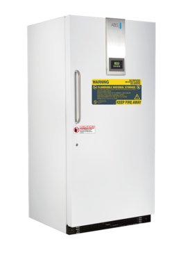 A tall, white laboratory freezer stands upright with a silver control panel near the top. It has a long vertical handle and warning labels on the door, including a large yellow label for flammable materials and a red caution sign below it.