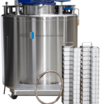 A large stainless steel cryogenic storage tank from American BioTech Supply stands beside two tall metal racks filled with white boxes. A metal circular lid or ring is placed in front of the tank. The tank has a blue digital control panel on top.