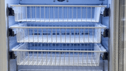 A refrigerated display case with three empty white wire shelves and a lit interior. The door is open, revealing the clean, blue-lit shelves, ready to store food or drinks. The refrigeration vents and temperature dial are visible at the back.